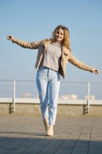 A young woman stands on one foot with arms outstretched, smiling under a clear blue sky. She wears