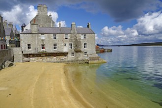 Old stone cottage on a quiet beach with clear water and partly cloudy skies, Lerwick, Shetland
