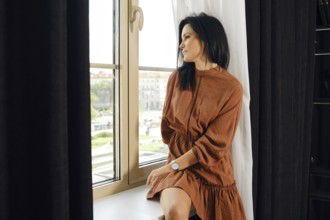 A woman in a brown dress relaxes by a large window, gazing thoughtfully outside. Sunlight filters
