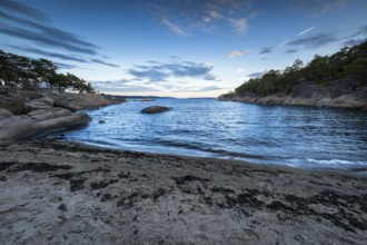 Small bay with sand and rocks, surrounded by pine trees under a partly cloudy sky, Skjeberg,