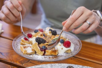 Hands holding spoons over waffles with cream and berries on a table