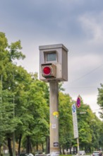 Traffic surveillance camera on a pole, surrounded by trees and road signs under a cloudy sky,