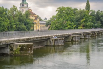 View of a bridge over a quiet river, surrounded by green trees and an impressive building in the