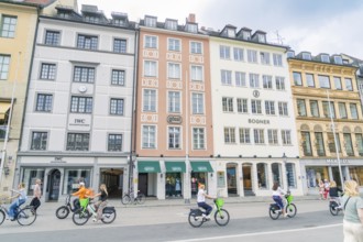 Passers-by ride along on bicycles in front of colourful building facades in Munich, Munich, Germany
