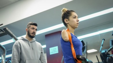A young female athlete with limb loss trains in a fitness gym with the support of her coach,