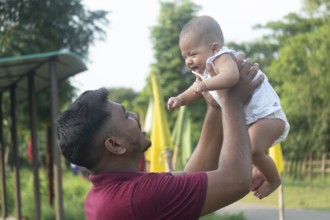 A man joyfully lifts a smiling baby in an outdoor setting-Dhaka, Bangladesh