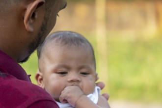 Baby calmly looks down, held by a gentle man outdoors-Dhaka, Bangladesh