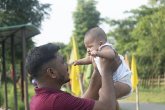 A parent is lifting a baby, creating a joyful moment in a green outdoor setting-Dhaka, Bangladesh