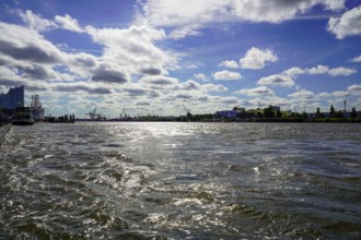 The Norderelbe with wavy water under a sunny sky, with urban background and silhouettes of cranes.
