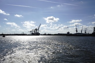 An industrial harbour with cranes and reflecting sunlight on the water under a blue sky. The view