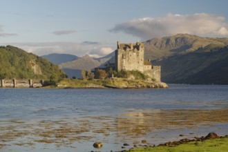 A picturesque castle on an island in the water, surrounded by mountains and a slightly cloudy sky,