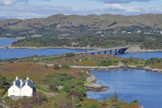 A white house set in a picturesque landscape of bridge, loch and rolling hills under a blue sky,