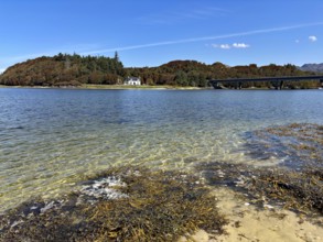 View over a loch with bridge and house surrounded by wooded hills under a blue sky, Silver Sands of