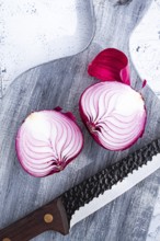 A vibrant red onion is cut in half, on a rustic wooden chopping board beside a sharp knife, ready