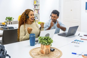Two diverse call center agents wearing headsets and working on laptops, celebrating their