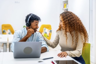 Two diverse colleagues in a modern office setup, a man wearing a headset using a laptop while a