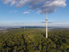 Television tower rises out of lush forest, surrounded by blue sky and decorated with clouds,
