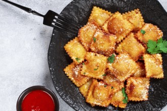 Fried ravioli with parmesan cheese, served with red sauce and garnished with herbs, on a black