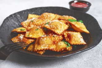 Fried ravioli with parmesan cheese, served with red sauce and garnished with herbs, on a black