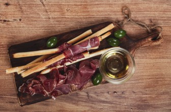 An appetizer of prosciutto, grissini and olives, served on a wooden board, with olive oil
