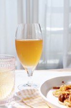 A glass filled with a light amber drink sits beside a plate of pasta. Bright sunlight streams
