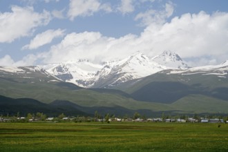 Green meadows in front of snow-covered mountains under a cloudy blue sky, landscape near Hartavan