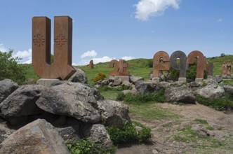 Row of stone columns with motifs, integrated into a rocky, green landscape under a blue sky,