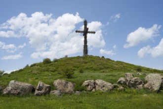 Metal cross on a green hill under a blue sky, surrounded by large stones, Monument to the Holy