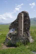 Stone with cross relief in front of a vast green landscape and blue sky, at the Monument of the