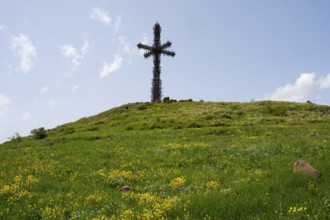 Metal cross on a green hill under a cloudy blue sky, surrounded by grass and yellow flowers,