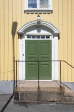 Entrance of a light yellow wooden house with a green door and white door frame, Fjällbacka,