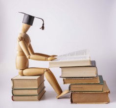 A wooden man in a student cap sits on a stack of books and reads a textbook