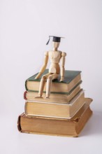 A wooden man in a student cap sits on a stack of textbooks on a white background