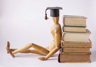 A wooden man in a student cap sits on the floor and leans his back on a stack of textbooks