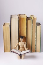 A wooden man in a student cap sits against a background of a stack of textbooks and reads a book,