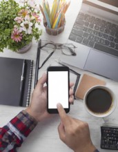 A person is using a cell phone on a desk with a laptop and a cup of coffee. The desk is cluttered