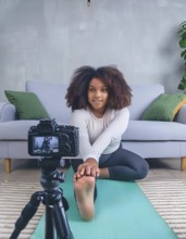 A young woman is doing a yoga pose in front of a camera. The camera is on a tripod and is recording