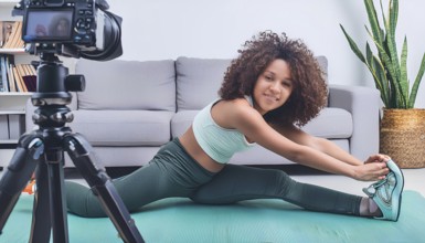 A young woman is stretching on a mat in front of a camera. The camera is on a tripod and is