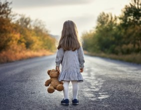 A young child stands alone holding a teddy bear in an urban outdoor setting, symbolizing innocence