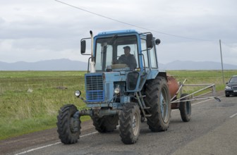 A blue tractor is driven by a man on a country road with fields and cloudy sky, Belarus MTZ-82