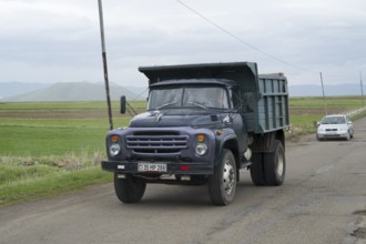 A blue lorry with a trailer drives on a country road surrounded by green fields and mountains,