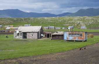 Rustic barns and buildings on green pasture against a mountainous backdrop with clouds in the sky,