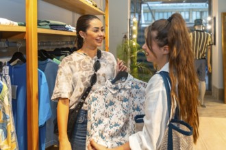 Two women interacting, choosing clothes in a store, one showing a shirt to the other, smiling and