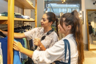 Two young women looking and choosing clothes in a fashion store, the shop assistant is helping a