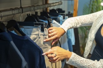 Female customer browsing through new collection in clothing store, selecting beige pants hanging on