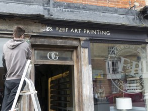 Decorator preparing wooden surface of shop front for painting, Devizes, Wiltshire, England, UK
