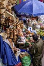 Traders at a lively market with ceramics, dolls and colourful fabrics under a blue umbrella,