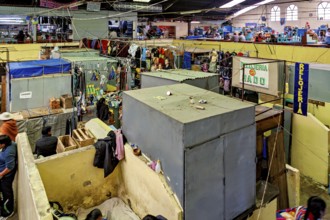 Lively market hall with numerous stalls and people in colourful clothing. Signs show various shops,