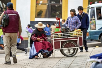 A saleswoman in a hat sits at a fruit stand on a busy pavement, selling fresh juice in the city of
