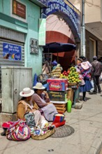 Women in traditional dress sell fruit at a busy market stall in a Bolivian town, selling goods in